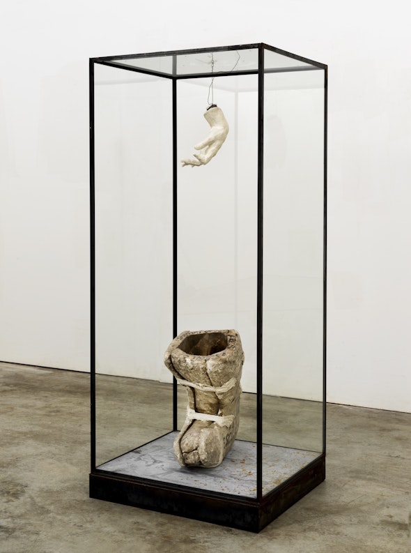 inside a tall vitrine by Kiefer, a plaster hand hangs from a wire. On the floor of the case is a plaster object in the shape of a boot.